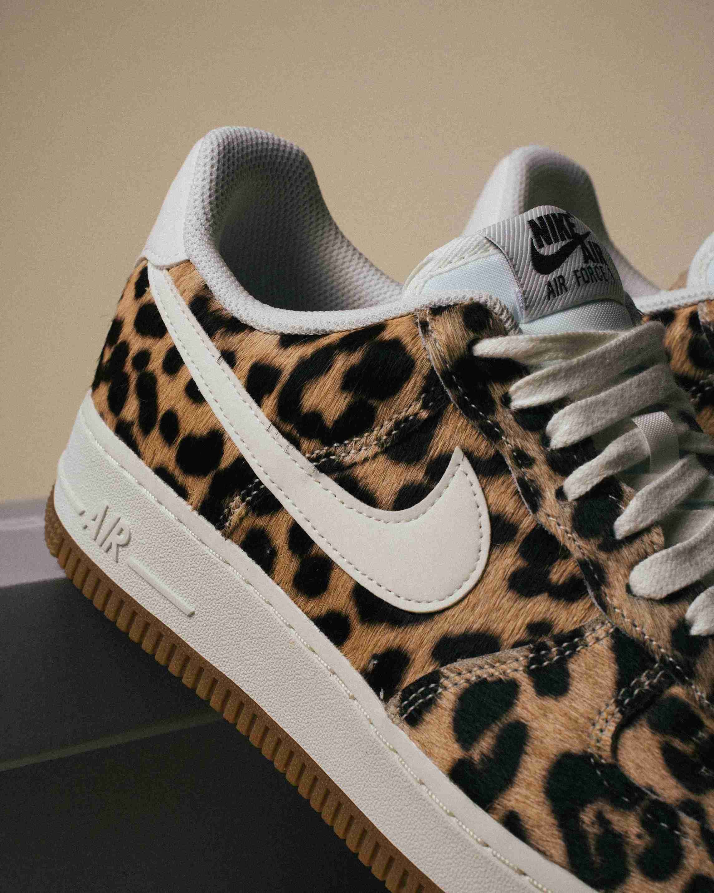 Paire de sneakers Nike Air Force 1 à motif léopard, placée à côté d’une boîte Nike grise sur un fond beige, vue artistique avec lumière douce.